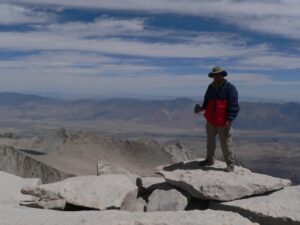 A man holding a GPS receiver stands at the peak of Mount Whitney