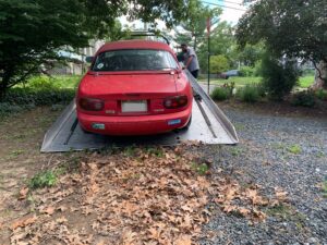 A red 1991 Mazda Miata with the hard top on is seen from the rear being loaded onto a flatbed trailer
