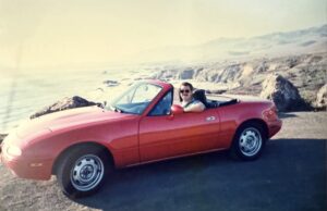 A red 1991 Mazda Miata convertible with the top down is parked at a turnout on the Pacific Coast Highway with the ocean and coastline in the background and a young man behind the wheel